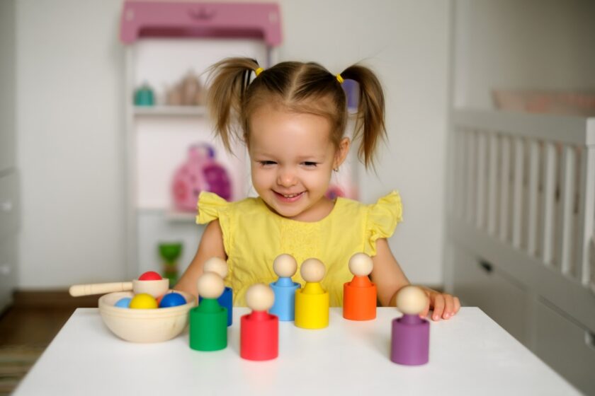 A young girl in a yellow dress smiles while playing with colorful wooden peg dolls and balls on a white table in a child’s room.