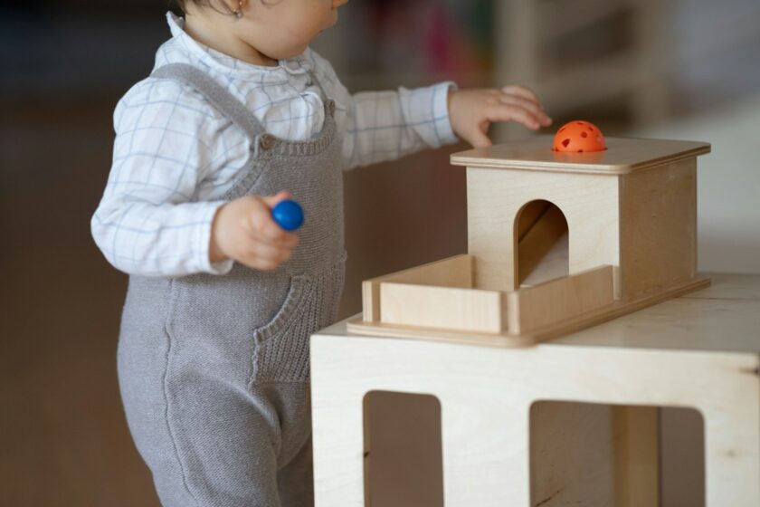 A toddler wearing grey overalls plays with a wooden toy, holding a blue ball and reaching toward an orange ball on top of the toy.